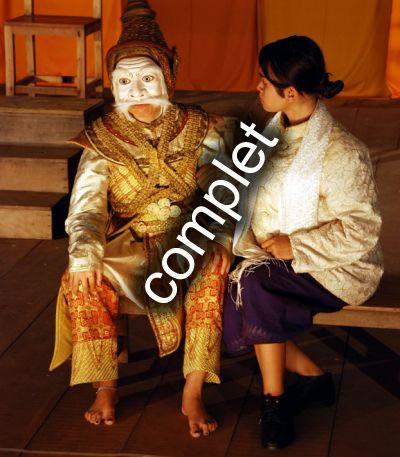 un personnage vêtu d'or porte un masque blanc, une jeune fille assise à ses cotés lui prend le bras