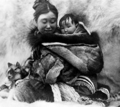 une femme inuit assise sur une peau de fourrure porte son bébé dans son dos. Elle sourit et le bébé bien emmitouflé dans une fourrure de phoque décorée regarde devant lui. Leurs visages sont proches. Contre elle, à sa droite un aperçoit le museau d'un chi